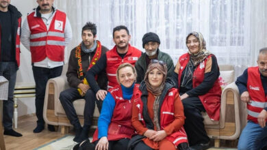 Bu görselde, bir evin oturma odasında çekilmiş samimi bir fotoğraf yer alıyor. Arka planda bir kanepeye oturmuş yaşlı bir adam, yanında bir kadın ve birkaç gönüllü bulunuyor. Gönüllüler kırmızı Türk Kızılayı yelekleri giymiş. Önde yere oturmuş iki kadın gönüllü var, arkada ise ayakta duran birkaç kişi görülüyor. Odanın zemini halı kaplı, arkada tül perdeli büyük bir pencere bulunuyor.