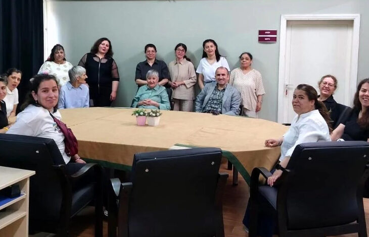 Görselde, büyük bir masa etrafında toplanmış çeşitli yaş gruplarından kadın ve erkekler görülüyor. Bazıları otururken bazıları ayakta duruyor ve kamera karşısında gülümsüyorlar. Masanın üzerinde süs bitkileri yer almakta. Arka planda açık renk duvar, kapı ve üzerinde "ARDUÇ" ve "LADİN" yazılı tabelalar bulunuyor. Bu görüntü, bir bakım merkezi veya sosyal hizmet kuruluşunda gerçekleştirilen bir ziyareti yansıtıyor olabilir.