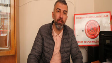 Görselde, koyu renk şişme mont ve açık pembe bir tişört giyen bir adam masa başında oturuyor. Sakallı ve hafif gri saçlı olan adam, doğrudan kameraya bakıyor. Arka planda "Eskişehir Bedensel Engelliler Sanat ve Spor Kulübü" yazılı kırmızı bir tabela bulunuyor. Masa üzerinde bir yazıcı veya fotokopi makinesi yer alıyor.