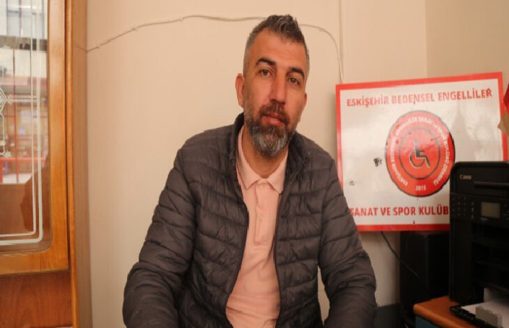 Görselde, koyu renk şişme mont ve açık pembe bir tişört giyen bir adam masa başında oturuyor. Sakallı ve hafif gri saçlı olan adam, doğrudan kameraya bakıyor. Arka planda "Eskişehir Bedensel Engelliler Sanat ve Spor Kulübü" yazılı kırmızı bir tabela bulunuyor. Masa üzerinde bir yazıcı veya fotokopi makinesi yer alıyor.