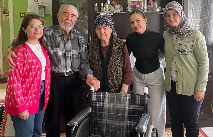 Görselde, bir ev ortamında beş kişi kameraya poz veriyor. Ortada yaşlı bir kadın ve yaşlı bir adam yer alıyor; kadın başörtüsü takıyor ve adamın koluna tutunmuş. Yaşlı çiftin yanında üç genç kadın bulunuyor; biri pembe hırka giymiş, biri siyah üst ve gri eşofman giymiş, diğeri ise başörtülü ve yeşil gömlekli. Önde bir tekerlekli sandalye görülüyor. Görsel, kuşaklar arası dayanışmayı, ziyaret ve destek temalı sıcak bir anı yansıtıyor.
