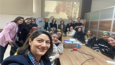 Görselde, bir toplantı ya da eğitim etkinliğinde bir araya gelmiş çoğunluğu kadınlardan oluşan bir grup görülüyor. Katılımcılar, uzun bir masa etrafında toplanmış ve ön planda yer alan bir kadın selfie çekiyor. Arkada bir projeksiyon ekranında bir erkeğin görüntüsü yansıtılmış; bu kişinin çevrim içi olarak etkinliğe katıldığı anlaşılıyor. Oda, modern ve aydınlık bir toplantı salonunu andırıyor. Katılımcıların gülümsemeleri ve enerjik halleri, etkinliğin pozitif ve destekleyici bir atmosferde geçtiğini yansıtıyor.