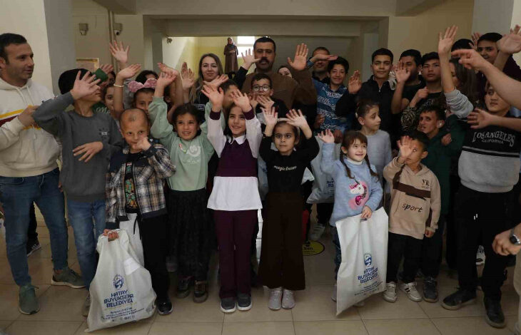 Görselde, çoğunluğu çocuklardan oluşan bir grup insan neşeyle ellerini havaya kaldırarak poz veriyor. Çocukların ellerinde "Hatay Büyükşehir Belediyesi" logolu beyaz çantalar bulunuyor. Arka planda yetişkinler de yer alıyor. İç mekânda çekilmiş olan bu kare, belediyenin sosyal bir etkinliği veya yardımı kapsamında gerçekleştirilen bir buluşmayı yansıtıyor.
