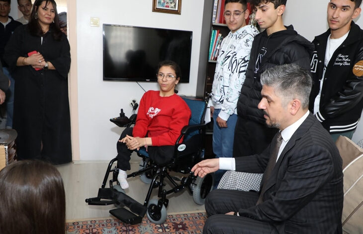 Görselde, kırmızı bir kazak giymiş genç bir kadın tekerlekli sandalyede oturuyor. Etrafında takım elbise giymiş bir adam ve birkaç genç erkek bulunuyor. Oda ev ortamında gibi görünüyor; duvarda bir televizyon, kitaplık ve halı mevcut. Arkada birkaç kişi daha izleyici konumunda duruyor. Görsel, sosyal bir ziyaret veya anlamlı bir yardım etkinliği sırasında çekilmiş izlenimi veriyor.