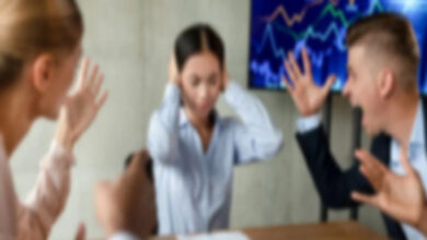 Görselde, bir toplantı odasında üç kişi yer almakta. Ortadaki kadın elleriyle kulaklarını kapatmış, üzgün ya da stresli görünüyor. Sağ ve sol taraftaki iki kişi ise ellerini kaldırmış ve yüksek sesle bağırıyor gibi görünüyor; bu da bir tartışma veya çatışma anını yansıtıyor. Arka planda, grafiklerin yer aldığı bir ekran bulunmakta. Görüntü bulanık olduğu için yüzler net seçilemiyor ancak ortamın gergin olduğu açıkça hissediliyor. Bu sahne, iş yerinde mobbing ya da stresli bir çalışma ortamını temsil ediyor olabilir.