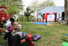 Fotoğrafta, açık havada düzenlenen bir etkinlik görülüyor. Çim alanda çocuklar sahnede gösteri yaparken, izleyiciler arasında tekerlekli sandalyede oturan bireyler de bulunuyor. Sahnenin arkasında Türk bayrağı, Atatürk posteri ve “ÖZGEM” yazılı bir afiş yer alıyor. Etkinlik alanı balonlar ve süslemelerle donatılmış. Bahar havası hâkim, ağaçlar yeşermiş. Aileler çocukları izlerken fotoğraf çekiyor, samimi ve neşeli bir atmosfer var.