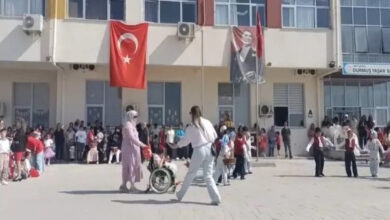 Bu görselde, bir okulun bahçesinde düzenlenen etkinlik görülüyor. Ön planda bir çocuk tekerlekli sandalyede, yanında ona eşlik eden bir yetişkin ve başka bir kişi yer almakta. Arka planda birçok çocuk ve yetişkin toplanmış, etkinliği izliyorlar. Binanın cephesinde Türk bayrağı ve Atatürk posteri asılı. Sağ üst köşede okulun adı “Durmuş Yaşar İlkokulu” olarak görünüyor.