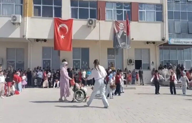 Bu görselde, bir okulun bahçesinde düzenlenen etkinlik görülüyor. Ön planda bir çocuk tekerlekli sandalyede, yanında ona eşlik eden bir yetişkin ve başka bir kişi yer almakta. Arka planda birçok çocuk ve yetişkin toplanmış, etkinliği izliyorlar. Binanın cephesinde Türk bayrağı ve Atatürk posteri asılı. Sağ üst köşede okulun adı “Durmuş Yaşar İlkokulu” olarak görünüyor.