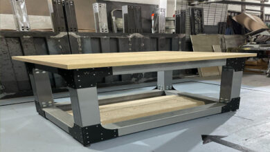 Bu görselde, endüstriyel tarzda tasarlanmış sağlam bir masa yer almakta. Masanın üst kısmı ahşap, ayakları ise metalden yapılmış; oldukça dayanıklı bir yapıya sahip olduğu görülüyor. Alt kısmında da destekleyici bir platform bulunmakta. Arka planda ise çeşitli metal parçalar, aletler ve sanayi tipi eşyalar yer almakta; bu da fotoğrafın bir atölyede çekildiğini gösteriyor.