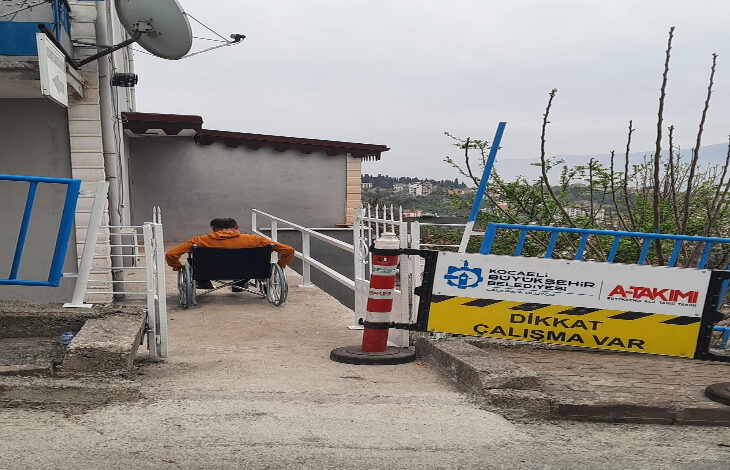 Görselde, turuncu mont giymiş bir kişi tekerlekli sandalyeyle bir rampada ilerliyor. Rampanın iki yanında beyaz metal korkuluklar bulunuyor. Sol tarafta bir bina, çanak anten ve mavi korkuluklar görülüyor. Sağ tarafta ise "Dikkat Çalışma Var" yazılı bir uyarı tabelası ve "Kocaeli Büyükşehir Belediyesi" logosu yer alıyor. Arka planda ağaçlar ve açık hava manzarası bulunuyor. Görüntü, engelliler için yapılan altyapı çalışmalarıyla ilgili olabilir.