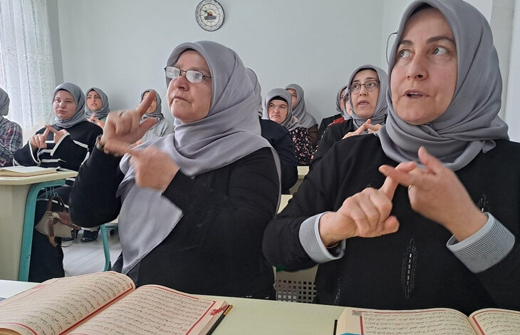Bu görselde, başörtülü kadınlardan oluşan bir grup bir sınıf ortamında oturuyor. Ön sıradaki iki kadın, elleriyle işaret dili kullanarak iletişim kuruyor. Masalarının üzerinde açık Kur'an-ı Kerimler bulunuyor. Arka planda da benzer şekilde Kur’an-ı Kerim okuyan ya da işaret diliyle öğrenmeye katılan diğer kadınlar yer almakta. Görseldeki atmosfer, dini eğitim alan işitme engellilere yönelik bir kursu yansıtıyor.