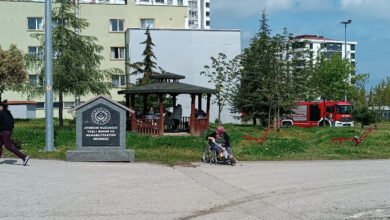 Bu görselde, bir huzurevinin dış cephesi ve çevresi görülmekte. Fotoğrafın sol ön planında "Atakum Huzurevi Yaşlı Bakım ve Rehabilitasyon Merkezi" yazılı bir tabela yer almakta. Arka planda bir kamelya içinde bazı insanlar oturuyor. Sağ tarafta bir kişi tekerlekli sandalyede oturuyor ve yanında ona yardım eden başka bir kişi bulunuyor. Arkada, yangın müdahalesi için hazır bekleyen bir itfaiye aracı dikkat çekiyor. Çevrede ağaçlar, çim alanlar ve banklar yer almakta.