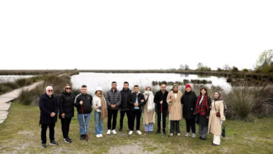 Bu görselde, doğayla iç içe bir alanda, su kenarında poz veren bir grup insan yer almakta. Grup, çeşitli yaş ve cinsiyetlerden bireylerden oluşuyor. Aralarından bazıları beyaz baston kullanıyor; bu da görme engelli bireylerin de grupta yer aldığını gösteriyor. Havanın kapalı olduğu bir günde, doğal bir ortamda çekilmiş bu fotoğrafta insanlar gülümseyerek kameraya poz veriyor.