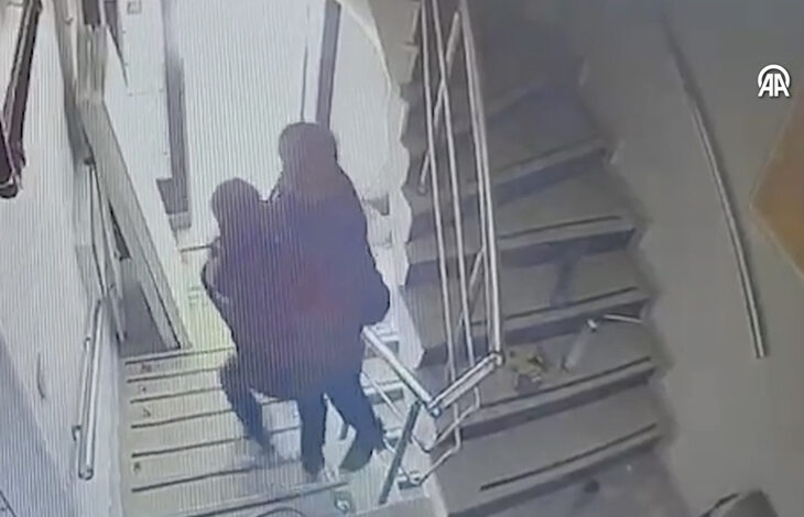 Görsel, bir güvenlik kamerası kaydını andıran, bulanık ve gri tonlarında bir sahneyi göstermektedir. Bir merdiven boşluğunda, iki kişi görülmektedir. Bir kişi, diğerini kollarının arasına almış ve yukarı doğru taşımaktadır. Taşıyan kişi koyu renkli giysiler giymektedir ve taşınan kişinin yüzü tam olarak seçilememektedir.