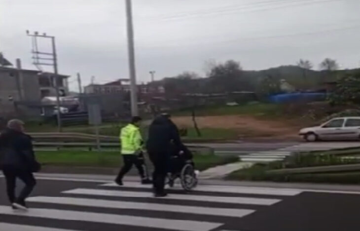 Görselde, bir yaya geçidinde tekerlekli sandalyedeki bir bireye yardımcı olan bir trafik polisi ve başka bir kişi görülüyor. Polis, reflektörlü sarı mont giymişken, diğer kişi koyu renk kıyafetler içinde. Arkada birkaç yaya daha geçidi kullanıyor. Çevrede kırsal bir alan, bazı binalar ve bir otomobil yer alıyor. Görsel, trafik yoğunluğu içinde engelli bireylere yardım edilmesini ve güvenli geçişin sağlanmasını yansıtan anlamlı bir anı gösteriyor.