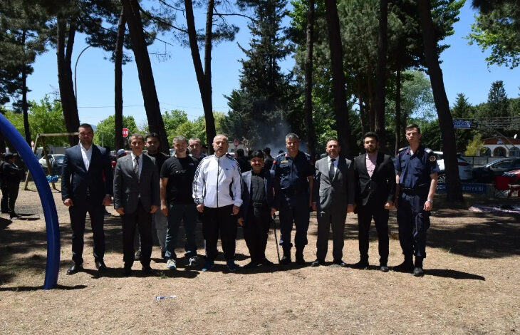 Bu görselde, 10-16 Mayıs Engelliler Haftası kapsamında Orhangazi’de düzenlenen bir etkinlikten kare yer alıyor. Açık havada, çam ağaçları arasında çekilmiş bu fotoğrafta, farklı kurum ve meslek gruplarından insanlar yan yana dizilmiş şekilde poz veriyor. Aralarında takım elbise giymiş kamu görevlileri, üniformalı emniyet mensupları ve sivil bireyler bulunuyor.