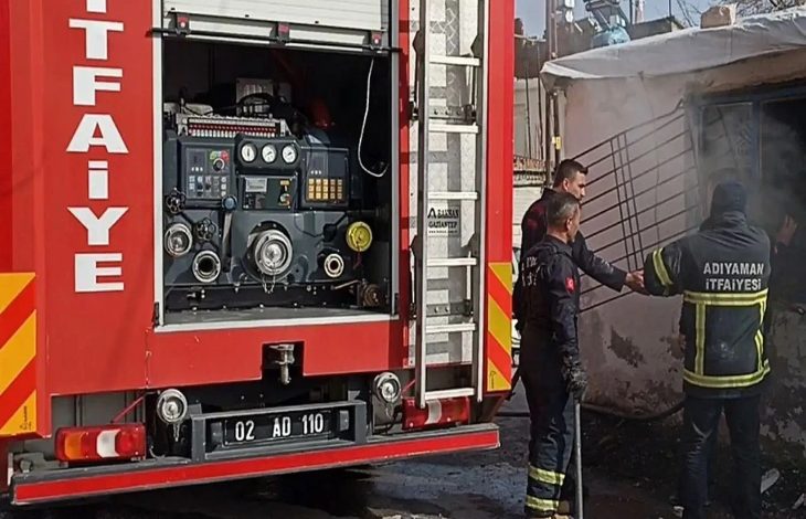 Bu karede Adıyaman İtfaiyesi ekiplerinin bir konteyner kentte çıkan yangına müdahale ettiği görülüyor. Görselin hemen sol tarafında itfaiye aracının arka bölümü, hortum ve kontrol paneli net biçimde seçiliyor. Sağda ise yangın çıkan yapıya yönelen birkaç itfaiye eri bulunuyor. Arka plandaki yoğun duman, olayın ciddiyetini gözler önüne seriyor.