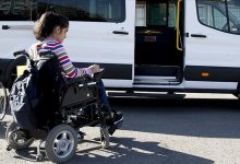 Görselde, tekerlekli sandalyede oturan genç bir kız, kapısı açık olan engelli erişimine uygun bir okul servisine binmek üzere. Servis aracı beyaz renkte ve yerde bir rampa sistemi görünmüyor ancak erişim sağlanacağı izlenimi veriliyor. Görsel, engelli bireylerin eğitime erişimini kolaylaştıran taşımalı eğitim uygulamasını yansıtıyor.