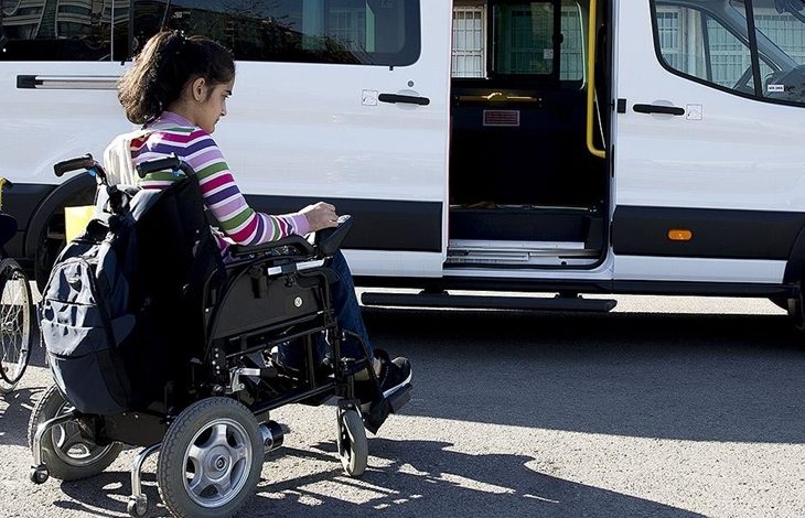 Görselde, tekerlekli sandalyede oturan genç bir kız, kapısı açık olan engelli erişimine uygun bir okul servisine binmek üzere. Servis aracı beyaz renkte ve yerde bir rampa sistemi görünmüyor ancak erişim sağlanacağı izlenimi veriliyor. Görsel, engelli bireylerin eğitime erişimini kolaylaştıran taşımalı eğitim uygulamasını yansıtıyor.
