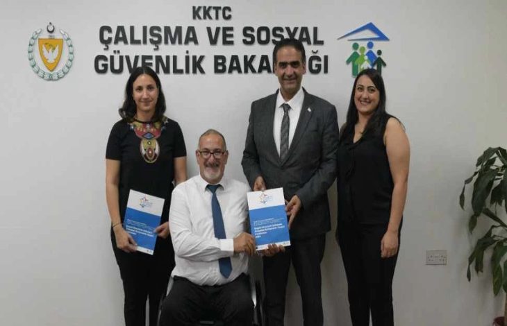 Görselde, KKTC Çalışma ve Sosyal Güvenlik Bakanlığı logosu önünde dört kişi poz vermektedir. Ortada oturan takım elbiseli ve kravatlı bir erkek elinde bir kitapçık tutarken, yanında ayakta duran diğer üç kişi de gülümsüyor. Arka planda bakanlığın adı Türkçe olarak duvarda yer almakta, sol üstte KKTC arması, sağ üstte ise insan figürlü bir logo görülmektedir.