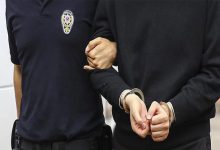 Görselde, bir polis memuru, elleri önden kelepçelenmiş bir kişiyi kolundan tutarak götürüyor. Polis memurunun üniformasında "Polis" yazısı ve amblemi net şekilde görülüyor. Fotoğraf, adli bir olay veya gözaltı durumunu temsil eden tipik bir kare. Kelepçelenmiş kişi sivil kıyafetli ve yüzü görünmüyor, mahremiyet korunmuş.