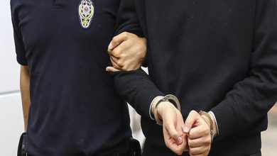 Görselde, bir polis memuru, elleri önden kelepçelenmiş bir kişiyi kolundan tutarak götürüyor. Polis memurunun üniformasında "Polis" yazısı ve amblemi net şekilde görülüyor. Fotoğraf, adli bir olay veya gözaltı durumunu temsil eden tipik bir kare. Kelepçelenmiş kişi sivil kıyafetli ve yüzü görünmüyor, mahremiyet korunmuş.