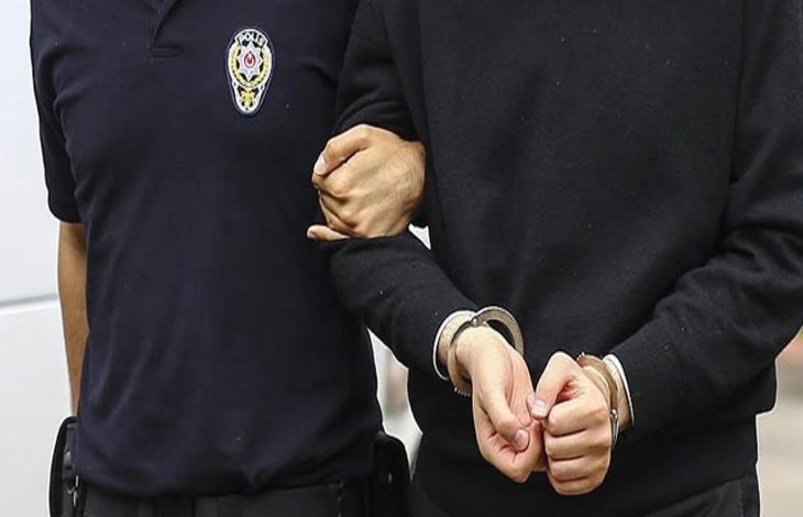 Görselde, bir polis memuru, elleri önden kelepçelenmiş bir kişiyi kolundan tutarak götürüyor. Polis memurunun üniformasında "Polis" yazısı ve amblemi net şekilde görülüyor. Fotoğraf, adli bir olay veya gözaltı durumunu temsil eden tipik bir kare. Kelepçelenmiş kişi sivil kıyafetli ve yüzü görünmüyor, mahremiyet korunmuş.