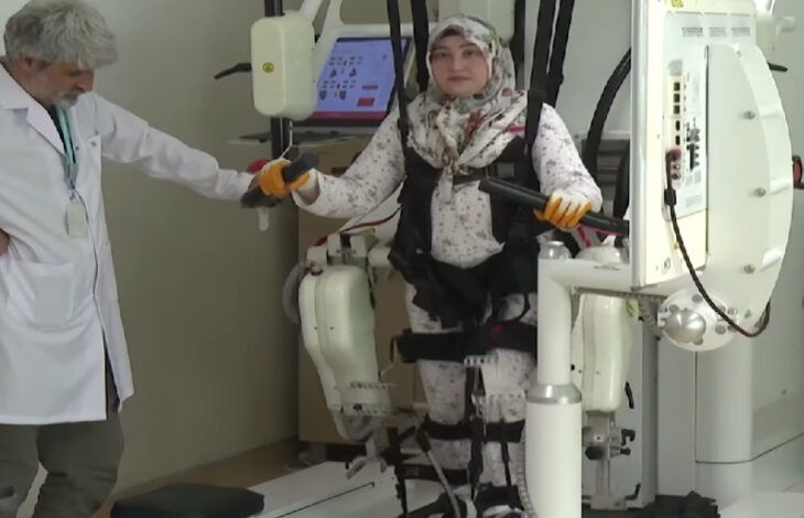 Görselde, bir kadın hasta bir yürüme robotu cihazına bağlı şekilde fizik tedavi görmekte. Cihaz, hastanın bacaklarına destek sağlayarak yürüme egzersizi yapmasına yardımcı oluyor. Kadının elleri tutacaklara bağlı, üzerinde emniyet kemerleri bulunuyor ve bir fizyoterapist ya da sağlık çalışanı da yanında destek veriyor. Arka planda tedavi sürecini izlemeye olanak tanıyan bir monitör yer alıyor.