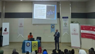 Bu görselde, bir konferans salonunda gerçekleşen bir sunum veya eğitim etkinliği görülüyor. Sahnenin ön kısmında biri masa başında oturan, diğeri ayakta sunum yapan iki kişi bulunuyor. Ayakta duran kişi mikrofonda bir şeyler anlatıyor, arka planda projeksiyonla yansıtılan bir sunum slaydı yer alıyor.