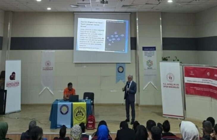 Bu görselde, bir konferans salonunda gerçekleşen bir sunum veya eğitim etkinliği görülüyor. Sahnenin ön kısmında biri masa başında oturan, diğeri ayakta sunum yapan iki kişi bulunuyor. Ayakta duran kişi mikrofonda bir şeyler anlatıyor, arka planda projeksiyonla yansıtılan bir sunum slaydı yer alıyor.
