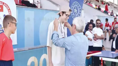 Bu görselde, bir spor etkinliği sırasında ödül töreni gerçekleşiyor. Önde, beyaz kıyafetler giymiş ve şapka takan genç bir sporcuya boynuna madalya takılırken görülüyor. Madalyayı, yaşça büyük bir kişi takıyor. Sporcu, gözlük takıyor ve gülümsüyor. Yanında kırmızı tişörtlü bir başka genç sporcu da bekliyor.