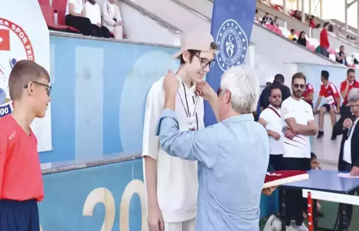 Bu görselde, bir spor etkinliği sırasında ödül töreni gerçekleşiyor. Önde, beyaz kıyafetler giymiş ve şapka takan genç bir sporcuya boynuna madalya takılırken görülüyor. Madalyayı, yaşça büyük bir kişi takıyor. Sporcu, gözlük takıyor ve gülümsüyor. Yanında kırmızı tişörtlü bir başka genç sporcu da bekliyor.