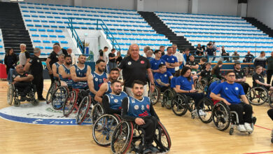 Bu görselde, bir spor salonunda düzenlenen ödül veya kutlama etkinliği sırasında çekilmiş bir an yer alıyor. Ön planda, tekerlekli sandalyedeki basketbol oyuncularından oluşan bir takım görülüyor. Sporcuların üzerinde koyu renkli formalar bulunmakta ve çoğunun yüzünde gurur ve mutluluk ifadesi var. Aralarında bir antrenör veya takım yetkilisi olduğu tahmin edilen, ayakta duran bir kişi de yer alıyor.