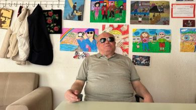 Bu fotoğrafta, gri tişört giymiş, gözlük takan bir görme engelli birey bir masanın başında oturuyor. Arka planda ise rengârenk çocuk resimleri ve çeşitli belgeler yer alıyor. Görsellerde sıkça empati, birliktelik ve erişilebilirlik temaları öne çıkıyor. Bazı çizimlerde baston kullanan bireyler, doğa manzaraları ve yardım temaları dikkat çekiyor.