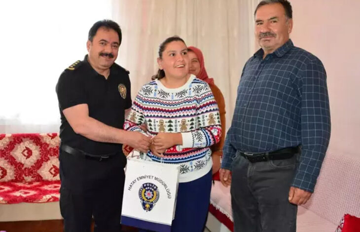 Görselde, bir ev ortamında çekilmiş samimi bir ziyaret anı yer almakta. Solda siyah üniformalı bir polis memuru (muhtemelen Hatay İl Emniyet Müdürü Kadir Gökce), ortada engelli bir genç kızla el sıkışıyor ve ona "Hatay Emniyet Müdürlüğü" logolu bir hediye çantası veriyor. Genç kız neşeli bir şekilde gülümsüyor. Sağda ise muhtemelen kızın babası olan bir adam duruyor; o da tebessüm ediyor. Arka planda bir kadın daha bulunmakta. Mekân sıcak ve sade bir oturma odası havasında.