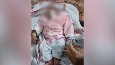 Bu görselde, bir bebek yatakta battaniyeye sarılmış şekilde yatıyor. Üzerinde pembe ve beyaz kıyafetler var. Burnundan veya ağızdan bir tıbbi tüp geçirilmiş ve bu tüp, yanında oturan bir yetişkinin elinde tuttuğu şırıngaya bağlı. Görsel, tıbbi desteğe ihtiyaç duyan bir bebeği ve ona bakım sağlayan bir yetişkini yansıtıyor.