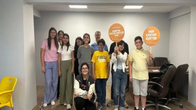 Görselde, bir toplantı odasında poz veren farklı yaşlardan çocuklar ve gençler yer alıyor. Ayakta duran bir grup bireyden bazıları ellerinde turuncu renkli dövizler tutuyor. Dövizlerde “HER ÇOCUK EŞİTTİR” ve “BİZİM DE ÇOCUK MECLİSİ VAR” ifadeleri yazıyor. Arka planda masa ve sandalyeler görülüyor. Görsel, İzmir Çocuk Meclisi'nin engelli hakları gündemiyle yaptığı toplantıya ait bir kareyi yansıtıyor.