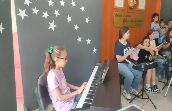 Görselde, genç bir kız dijital piyano çalarken görülüyor. Üzerinde mor bir tişört ve saçlarında yeşil toka olan bu kız, dikkatle tuşlara odaklanmış. Arka planda gitar çalan ve ritim tutan diğer öğrenciler yer alıyor. Duvarda beyaz yıldız figürleri ve Atatürk’ün bir sözüyle birlikte büstü dikkat çekiyor.