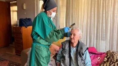 Fotoğrafta, yeşil önlük giymiş genç bir kadın, yaşlı bir kadının saçını tarak ile tutuyor ve kesim yapıyor.