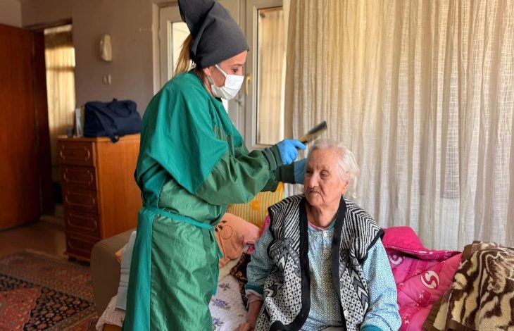Fotoğrafta, yeşil önlük giymiş genç bir kadın, yaşlı bir kadının saçını tarak ile tutuyor ve kesim yapıyor.