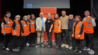 Görselde, sahne üzerinde bir grup insan poz vermiş durumda. Gruptakilerin bir kısmı turuncu reflektörlü yelekler giymiş; bu kişiler muhtemelen gönüllü ya da görevli. Ortada yer alan kişiler ise resmi kıyafetli ya da sivil giyimli. Arka planda “Afet Duyarlılığı” temalı bir afiş ve bir projeksiyon perdesi yer alıyor. Bu sahne, muhtemelen bir afet farkındalık etkinliği ya da seminerine ait.