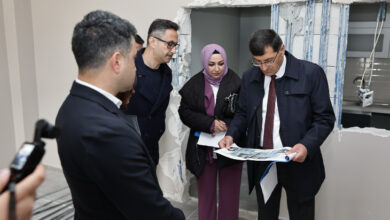 Bu görselde, dört kişilik bir grup bir binanın iç kısmında inceleme yaparken görülüyor. Arka planda, duvarın bir kısmı kırılmış ve içinden elektrik tesisat kabloları görünüyor; bu da yapının tadilat veya yenileme aşamasında olduğunu gösteriyor.