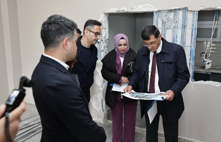 Bu görselde, dört kişilik bir grup bir binanın iç kısmında inceleme yaparken görülüyor. Arka planda, duvarın bir kısmı kırılmış ve içinden elektrik tesisat kabloları görünüyor; bu da yapının tadilat veya yenileme aşamasında olduğunu gösteriyor.