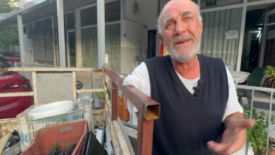 Bu görselde, Muğla’nın Marmaris ilçesinde yaşayan bir adam %71 engelli olduğu belirtilen Hüseyin İnce açık havada, bir tezgâh ya da taşıma aracının yanında görülüyor. Yaşlıca ve beyaz sakallı bu adam, üzgün veya çaresiz bir ifadeyle elini havaya kaldırmış, sanki bir durumu anlatıyor ya da şikâyet ediyor. Arka planda, apartman benzeri yapılar ve bazı eşyalar (saksılar, kaplar) dikkat çekiyor.