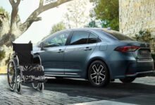 Görselde modern bir otomobilin (Toyota Corolla) hemen önünde duran boş bir tekerlekli sandalye yer almakta. Arka planda taş duvarlı bir ev ve doğa manzarası görülüyor. Fotoğraf, engelli bireylerin mobilite ihtiyaçlarını ve taşıma kolaylığını sembolize ediyor. Aynı zamanda, otomobilin engelli bireyler için önemli bir özgürlük ve bağımsızlık aracı olduğunu da ima ediyor.