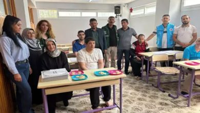 Bu görselde, bir sınıf ortamında bir grup insan yer alıyor. Önde, sıralarda oturan bireylerin çoğunluğunu görme engelli erkekler oluşturuyor. Masaların üzerinde renkli tabaklarda ikramlıklar yer almakta. Arkada ise çeşitli yaş gruplarından kadın ve erkeklerden oluşan destekleyici bir ekip ayakta poz veriyor. Mekân sade, halı kaplı ve aydınlık; eğitim ortamı olduğu hemen hissediliyor.