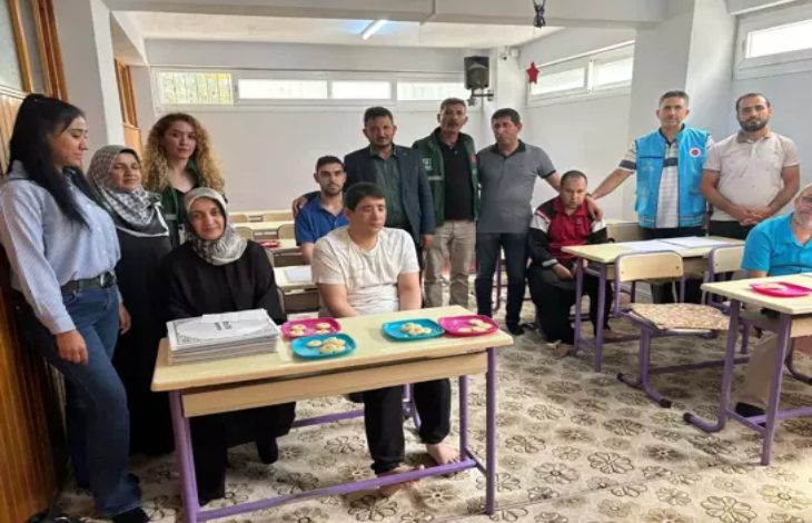 Bu görselde, bir sınıf ortamında bir grup insan yer alıyor. Önde, sıralarda oturan bireylerin çoğunluğunu görme engelli erkekler oluşturuyor. Masaların üzerinde renkli tabaklarda ikramlıklar yer almakta. Arkada ise çeşitli yaş gruplarından kadın ve erkeklerden oluşan destekleyici bir ekip ayakta poz veriyor. Mekân sade, halı kaplı ve aydınlık; eğitim ortamı olduğu hemen hissediliyor.