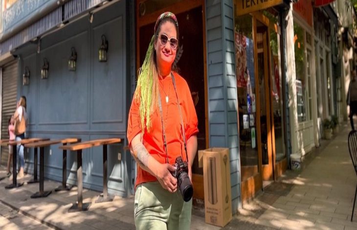 Görselde, bir kadın şehir sokaklarında poz veriyor. Üzerinde turuncu tişört, açık renk pantolon ve boynunda bir fotoğraf makinesi var. Uzun yeşil saçları dikkat çekici ve güneş gözlüğü takıyor. Arka planda kafe ya da restoranlar bulunuyor. Kadın özgüvenli bir duruşla objektife bakıyor.