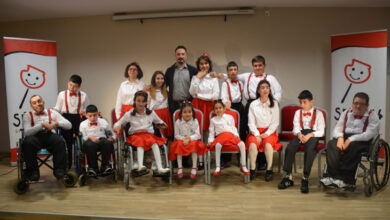 Bu görselde, sahnede toplu olarak poz veren bir grup çocuk ve genç yer alıyor. Katılımcıların çoğu tekerlekli sandalye kullanıyor ve hepsi uyumlu şekilde beyaz üst ve kırmızı alt kıyafetler giymiş. Arka planda ise Serebral Palsili Çocuklar Derneği'nin (SERÇEV) logosu yer alan iki pankart dikkat çekiyor.