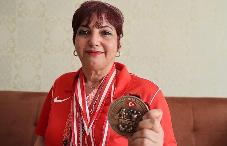 Görselde, kırmızı tişört giymiş orta yaşlarda bir kadın gülümseyerek elindeki madalyayı kameraya doğru tutuyor. Boynunda birden fazla madalya yer alıyor. Madalya üzerinde “Türkiye Vücut Geliştirme Fitness ve Bilek Güreşi Federasyonu” (TVGFBF) logosu görülüyor. Kadın, büyük olasılıkla bilek güreşi dalında başarı elde etmiş bir sporcu.